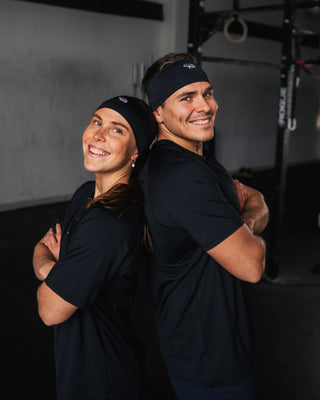 Two people wearing black athletic shirts and headbands standing back-to-back in a gym setting.